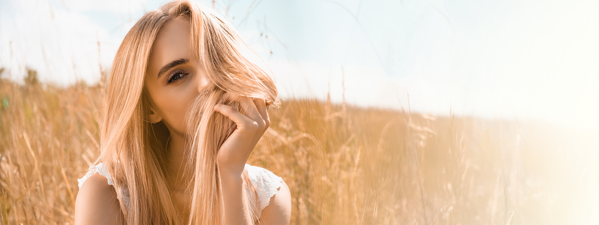 Blonde woman playfully holding her hair in a sunlit field, showcasing vibrant hair color and a natural, carefree atmosphere, relevant to Envy Studio's hair services and bridal offerings.