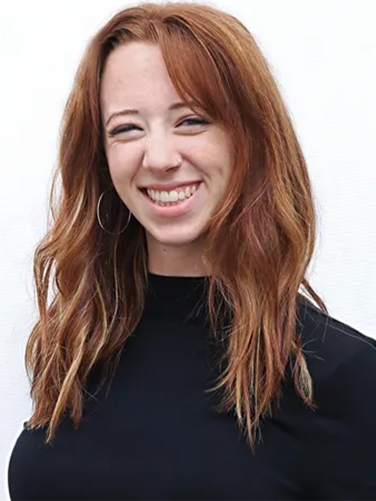 Abby, men's grooming specialist at Envy Studio, smiling with long, wavy red hair, wearing a black top, showcasing her expertise in haircuts and styling.