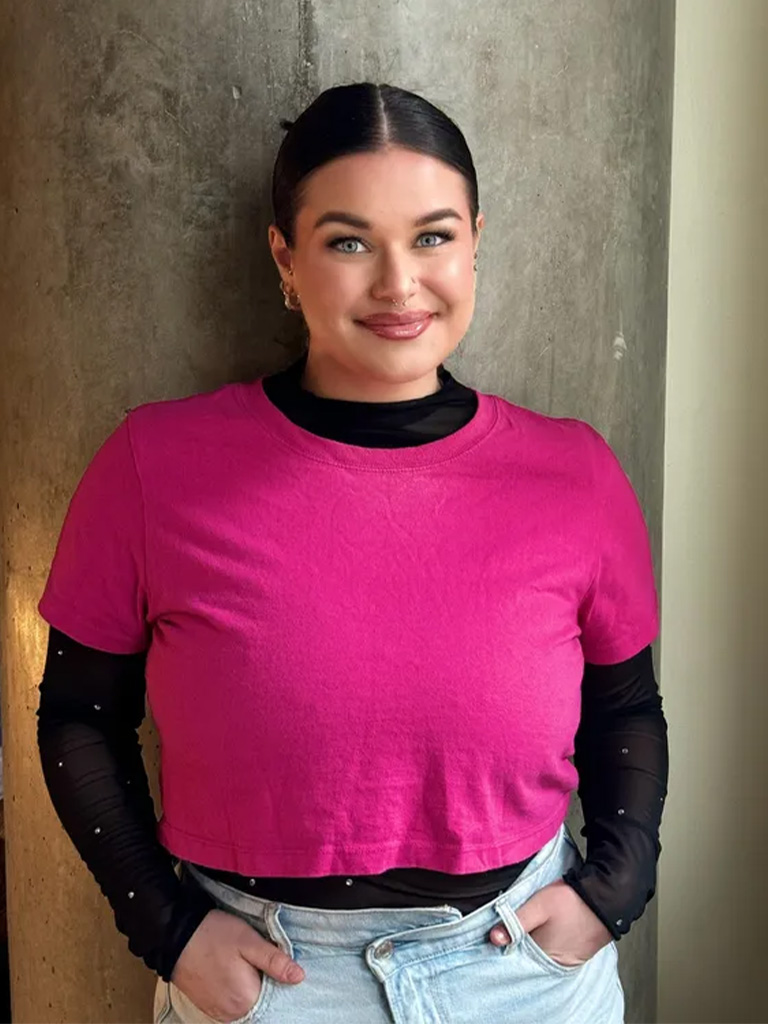 Lauren, lash artist at Envy Studio, wearing a pink cropped shirt and black long-sleeve top, smiling confidently against a textured wall.