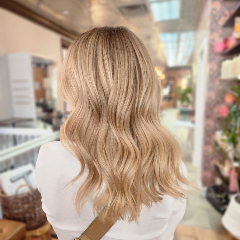 A person with wavy, shoulder-length blonde hair is seen from behind in a well-lit salon. | Envy Studio, Lancaster, PA