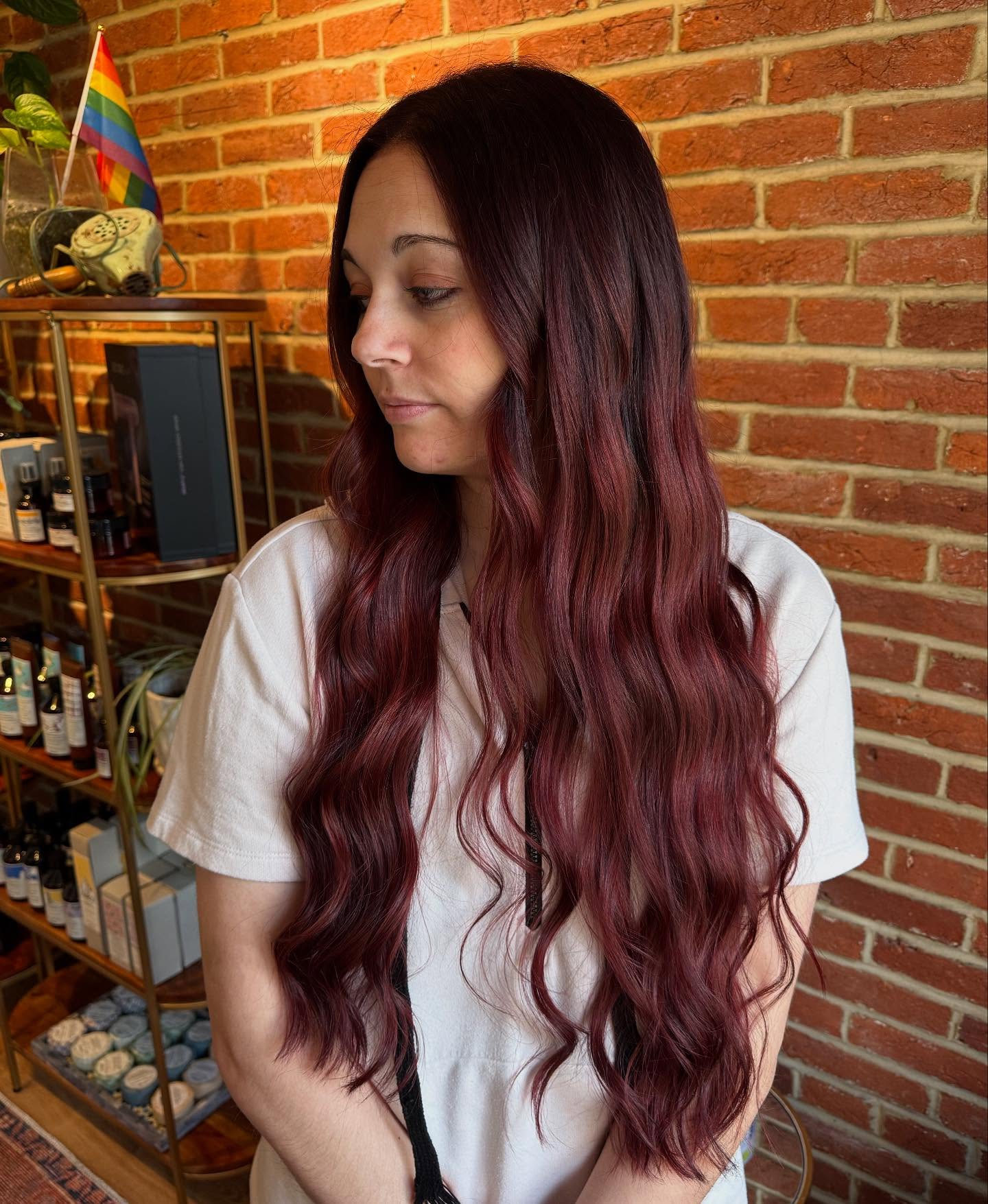 Woman with long, wavy dark red hair wearing a white shirt stands indoors near shelves and a brick wall, looking to the side. | Envy Studio, Lancaster, PA