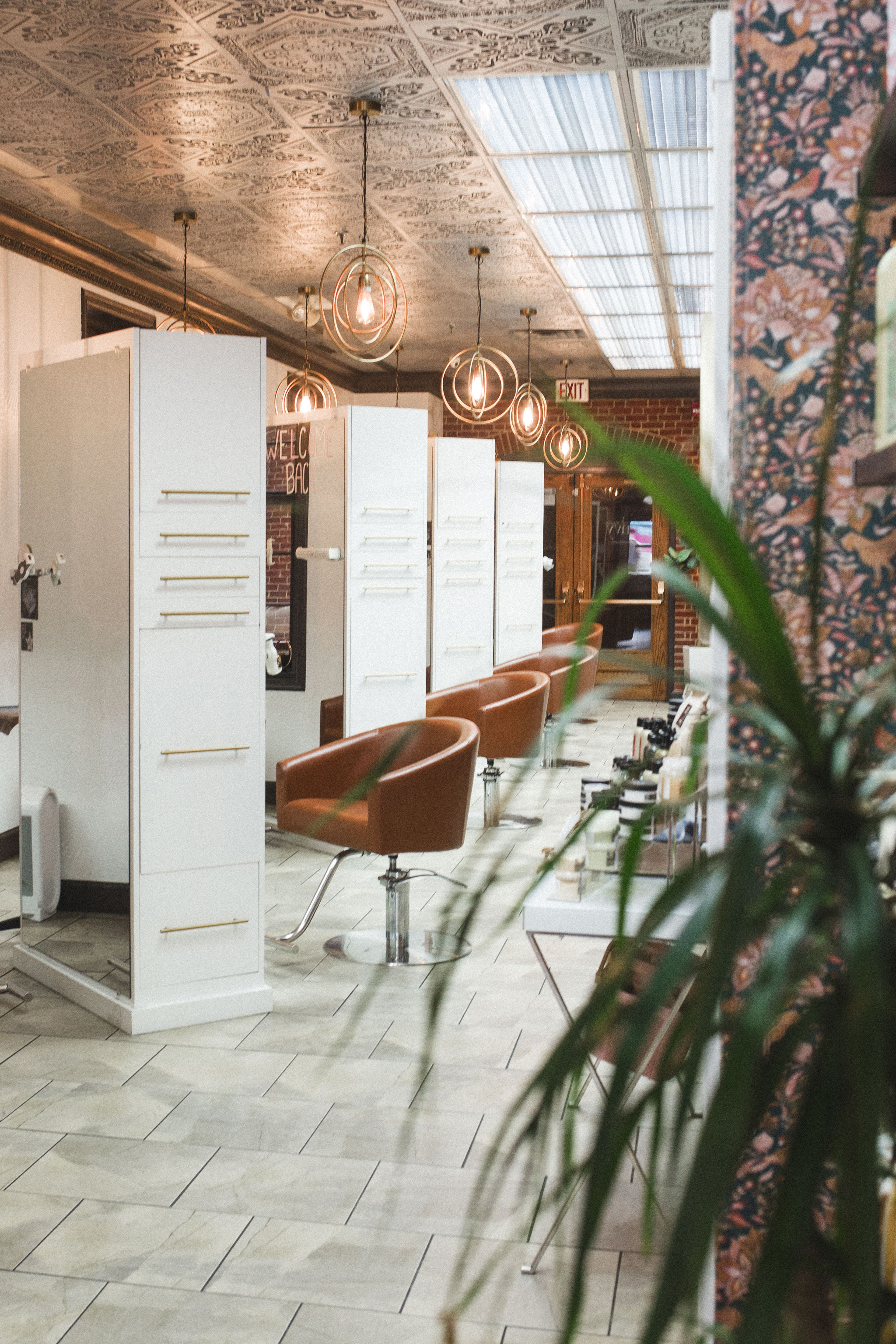 A modern hair salon with brown leather chairs, white styling stations, pendant lights, and tile flooring. A potted plant is visible in the foreground. | Envy Studio, Lancaster, PA