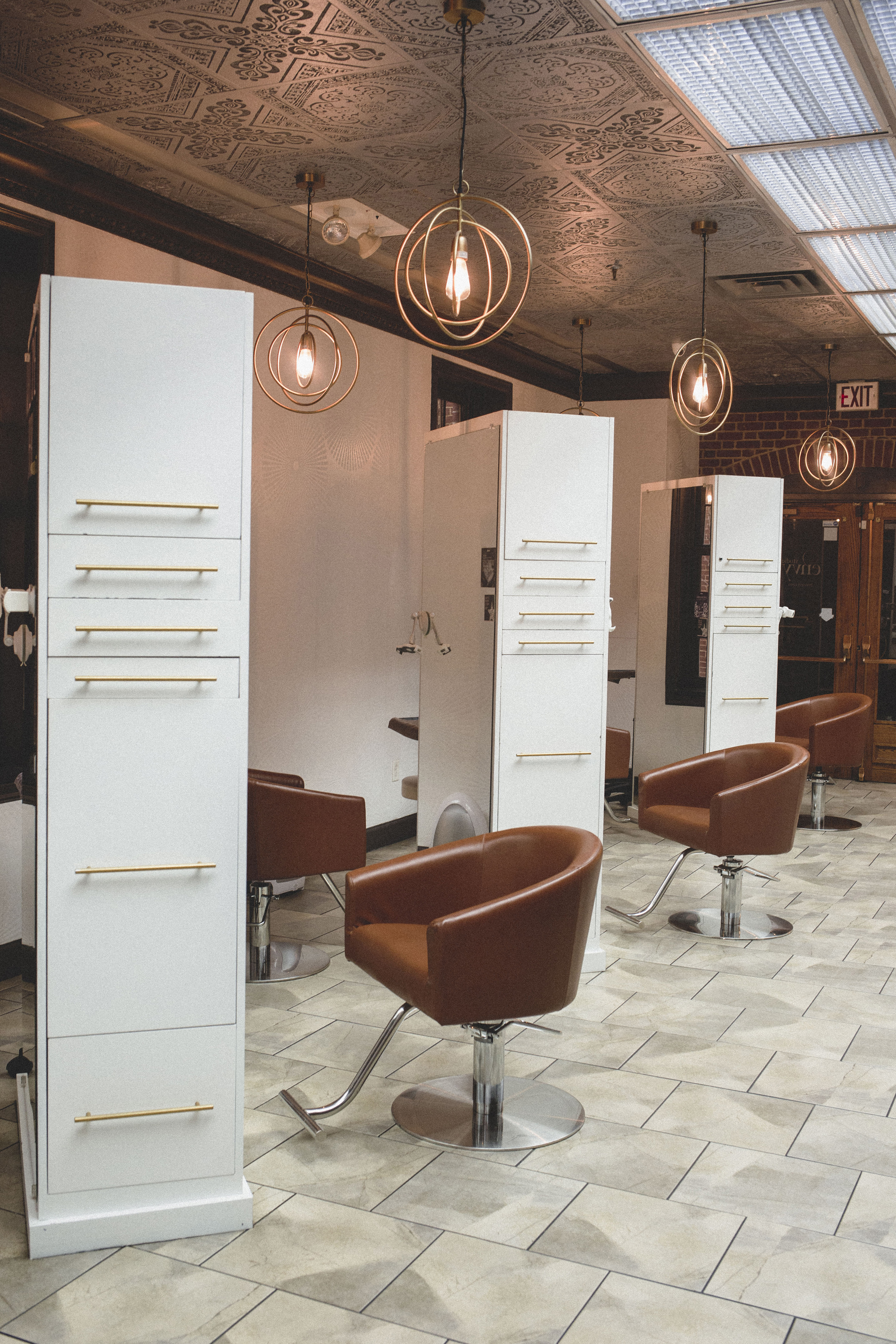 Modern hair salon interior with brown leather chairs, white styling stations, and hanging pendant lights, featuring tile flooring and decorative ceiling panels. | Envy Studio, Lancaster, PA