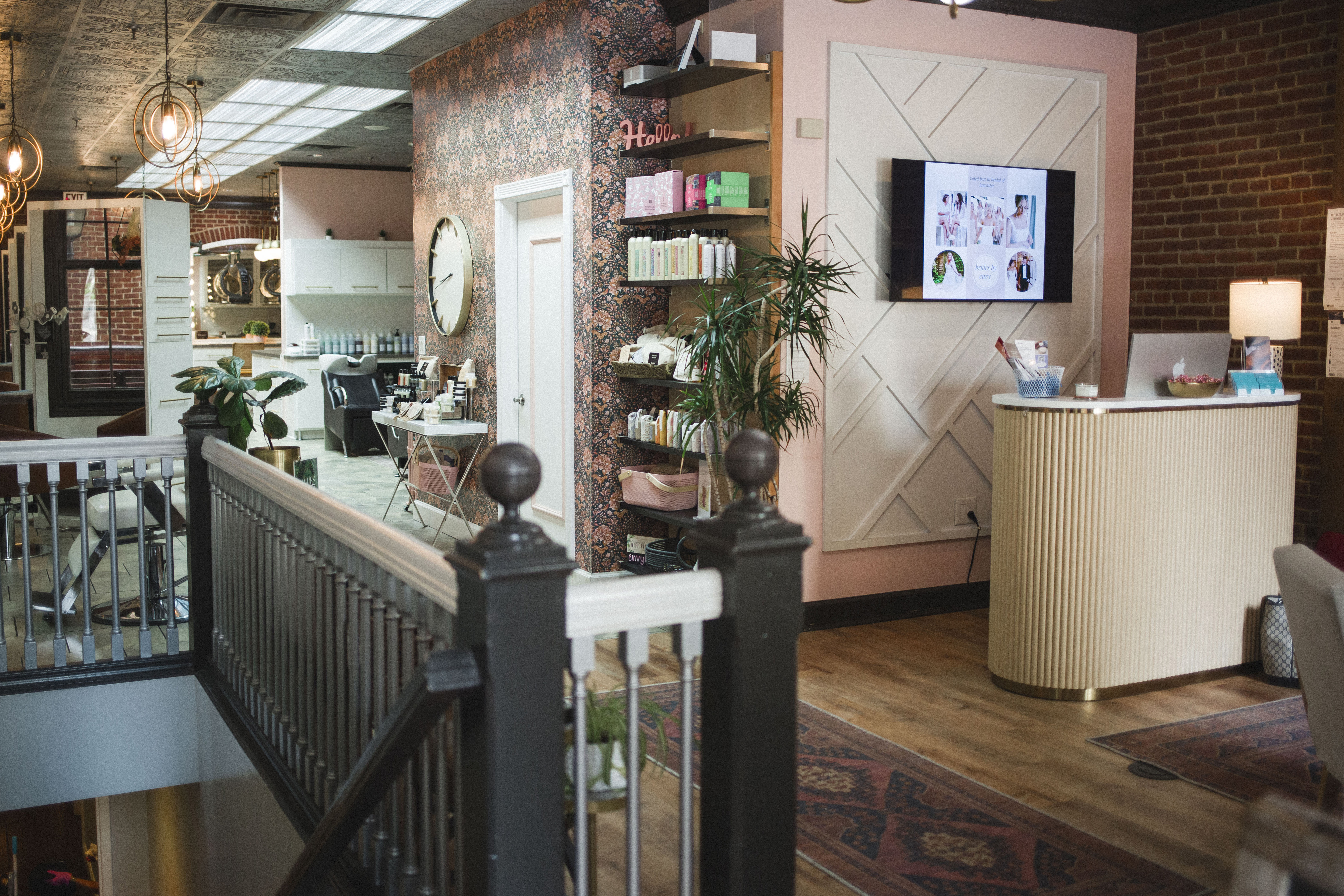 Modern salon interior with a reception desk, product shelves, a TV on the wall, plants, and salon stations visible in the background. Exposed brick and patterned wallpaper decorate the space. | Envy Studio, Lancaster, PA