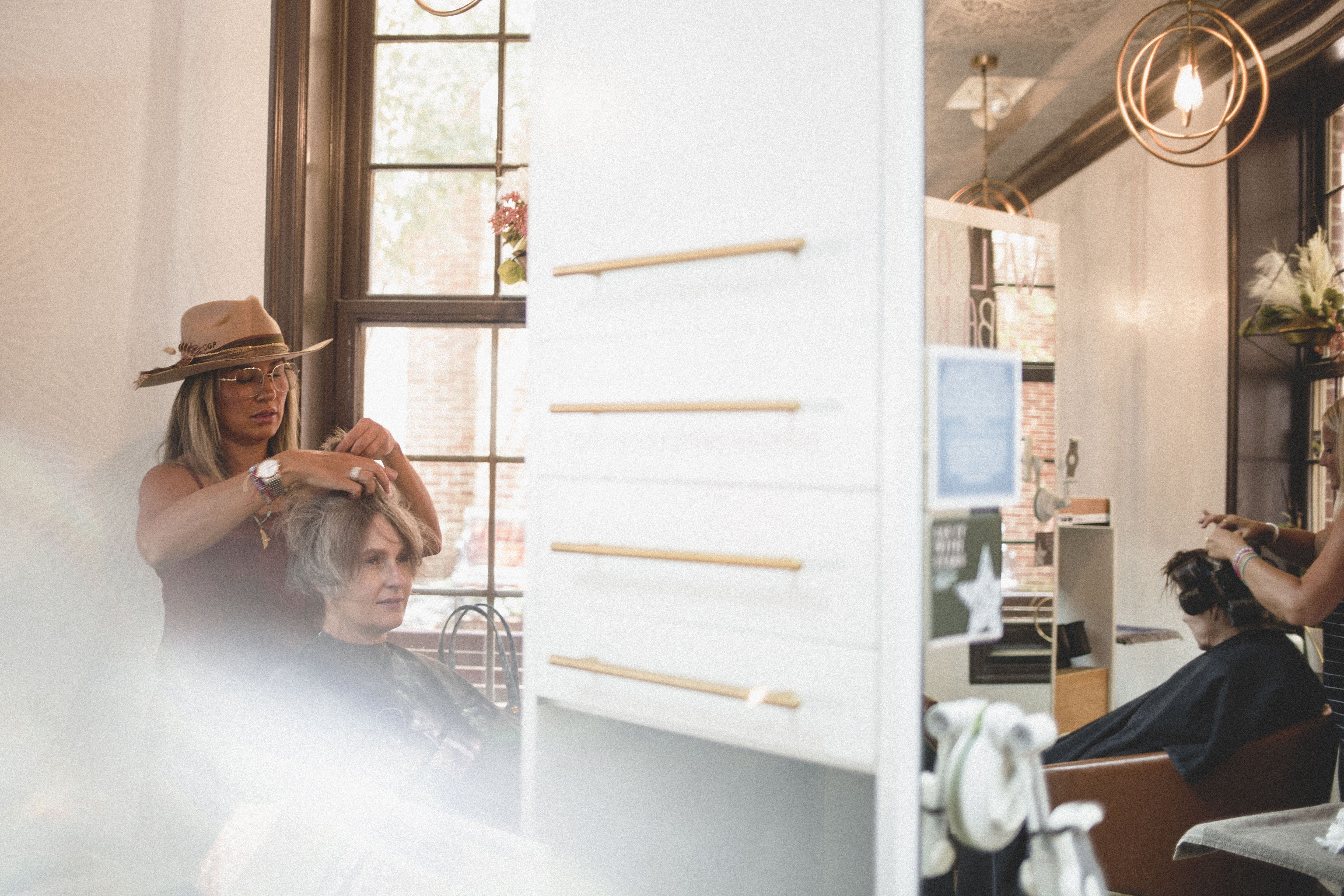 Two hairstylists work on clients’ hair inside a salon with large windows, wooden accents, and natural light streaming in. | Envy Studio, Lancaster, PA