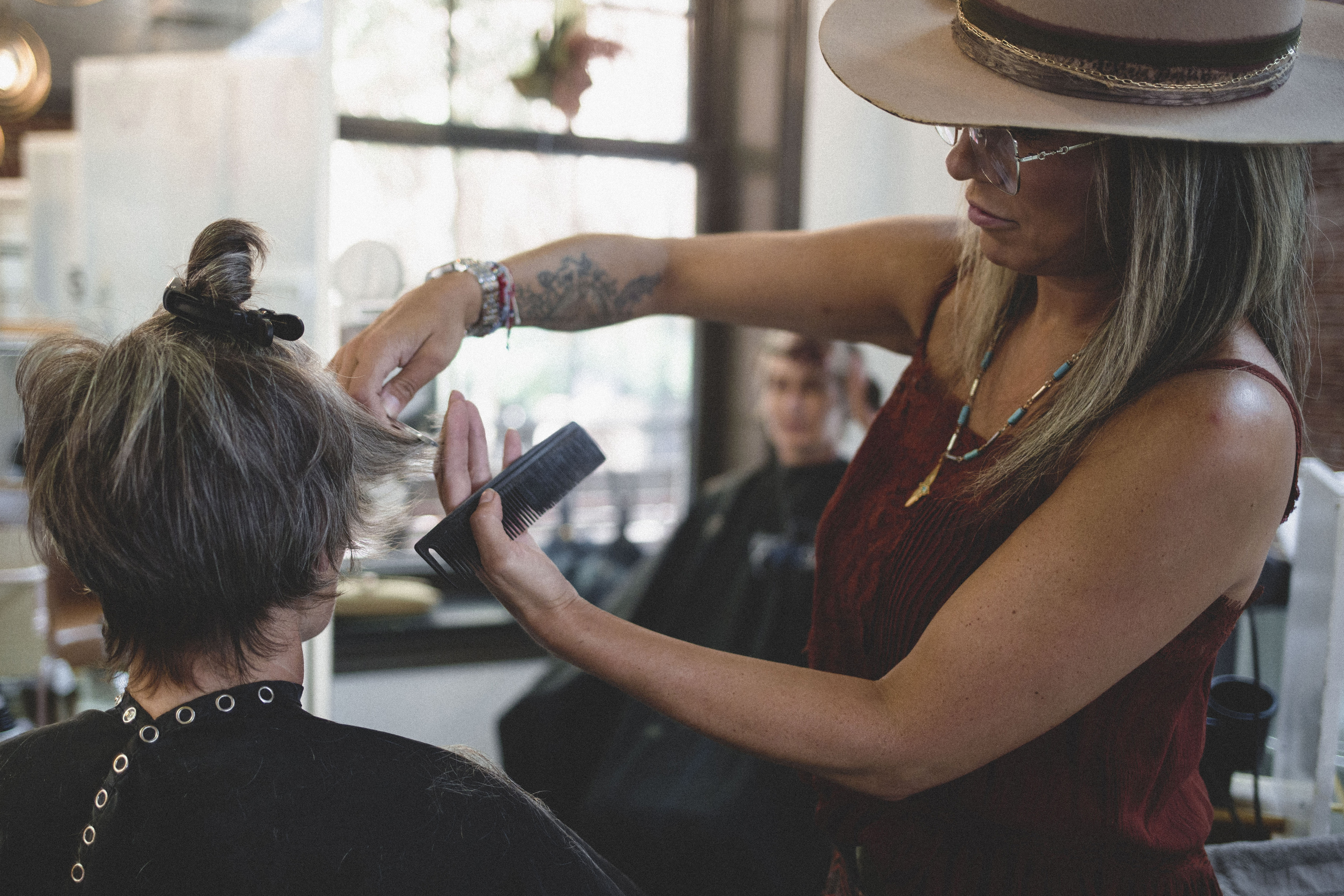 A hairstylist wearing a hat trims the hair of a seated client in a salon, holding scissors in one hand and a comb in the other. | Envy Studio, Lancaster, PA