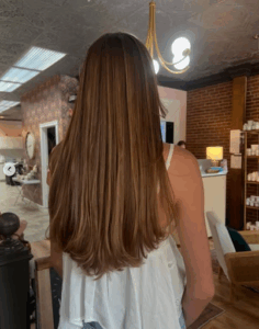 A person with long, straight, light brown hair stands indoors with their back to the camera in a well-lit, modern salon. | Envy Studio, Lancaster, PA