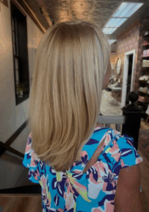 Woman with straight, shoulder-length blonde hair wearing a colorful floral top, standing indoors near a staircase in a well-lit room. | Envy Studio, Lancaster, PA