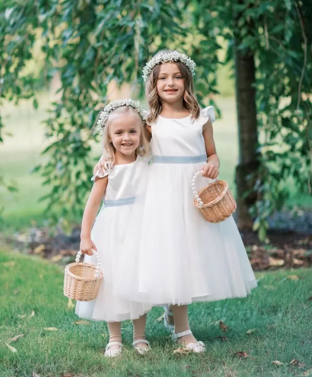 Two young girls in white dresses and flower crowns stand on grass, each holding a small wicker basket, with trees and greenery in the background. | Envy Studio, Lancaster, PA