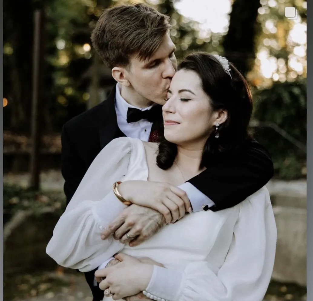 A man in a tuxedo embraces and kisses a woman in a white dress on the forehead outdoors, both appearing calm and content. | Envy Studio, Lancaster, PA