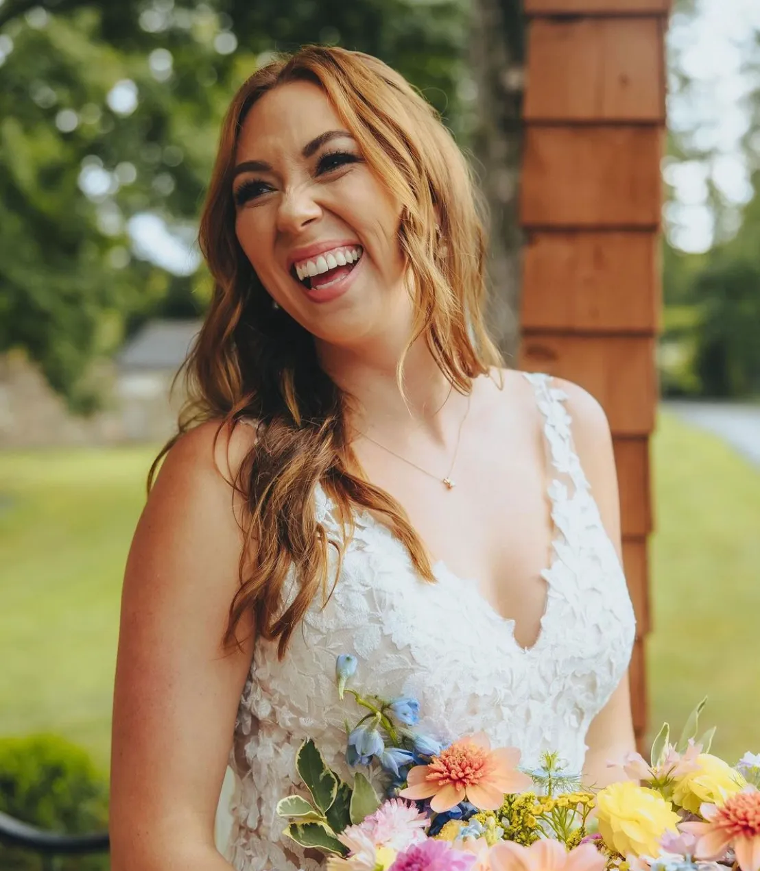A woman in a white lace dress smiles while holding a colorful bouquet of flowers, standing outdoors near a wooden structure. | Envy Studio, Lancaster, PA
