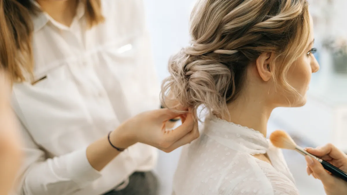 A person with blonde hair styled in an updo is having their hair adjusted while another person applies makeup to their face. | Envy Studio, Lancaster, PA