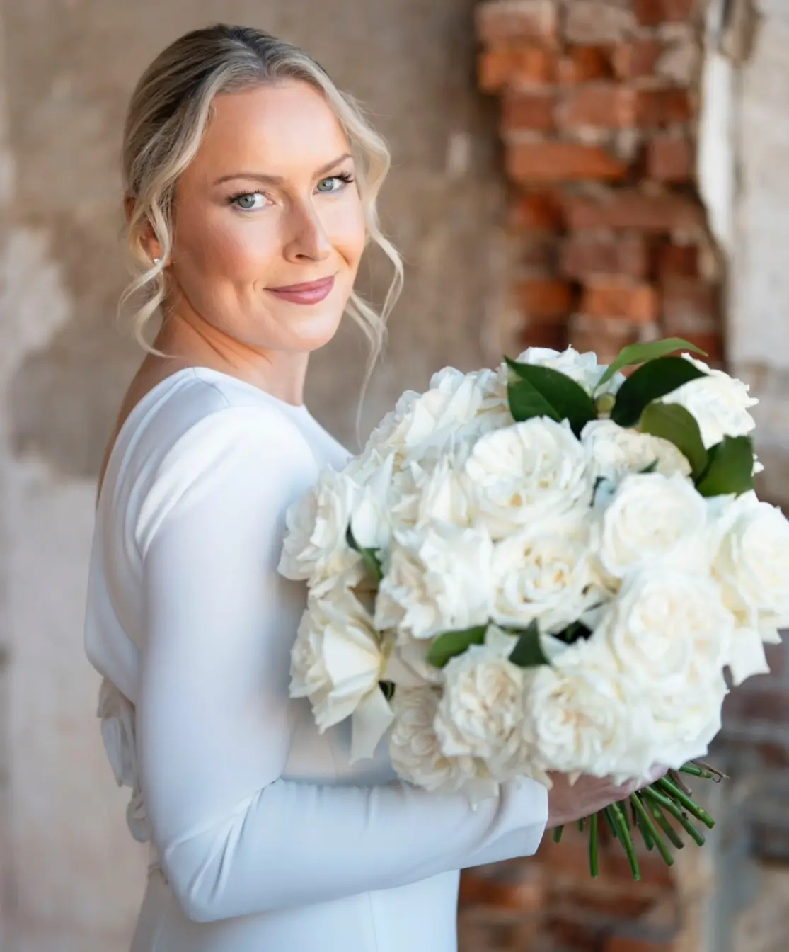 A woman in a white dress holds a large bouquet of white roses and greenery, standing in front of a textured brick and plaster wall. | Envy Studio, Lancaster, PA