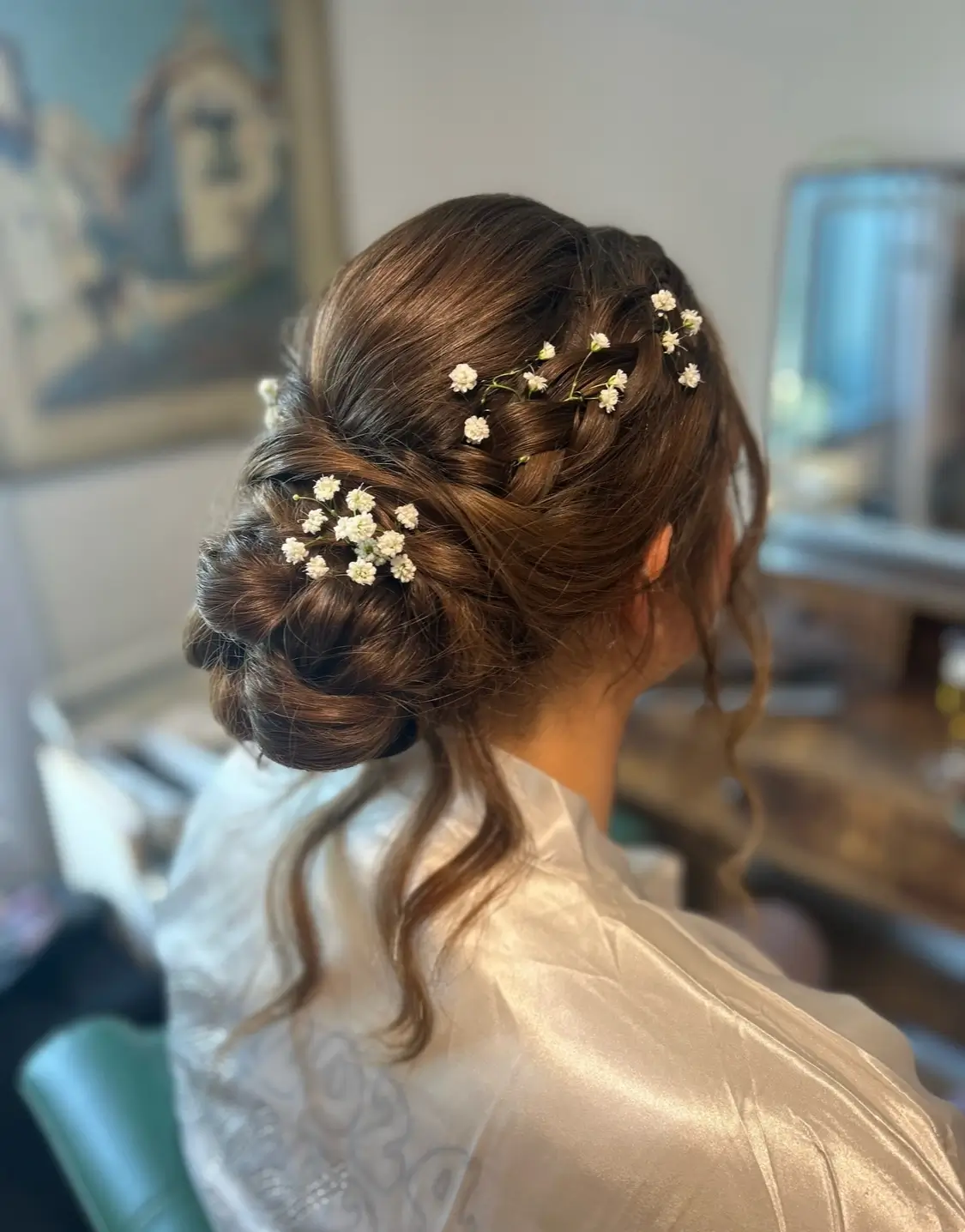 A woman with brown hair styled in a braided low bun, adorned with small white flowers, sits in front of a vanity mirror. | Envy Studio, Lancaster, PA