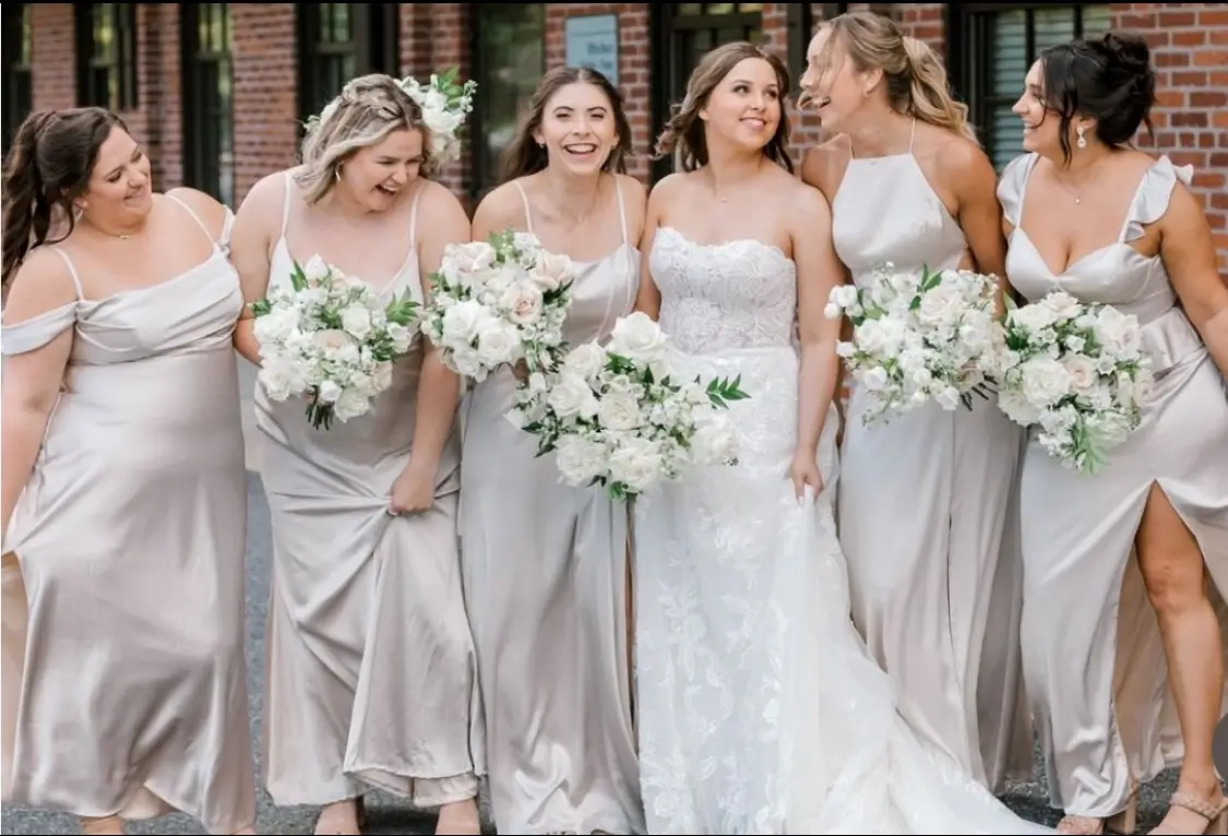 A bride in a white wedding dress stands outdoors with five bridesmaids in light-colored dresses, all holding white floral bouquets and smiling. | Envy Studio, Lancaster, PA