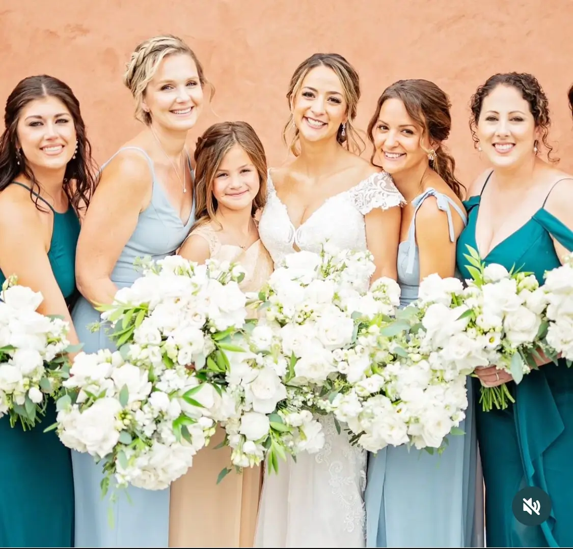 A bride and five bridesmaids, all holding large white flower bouquets, stand together smiling in front of a peach-colored wall. | Envy Studio, Lancaster, PA