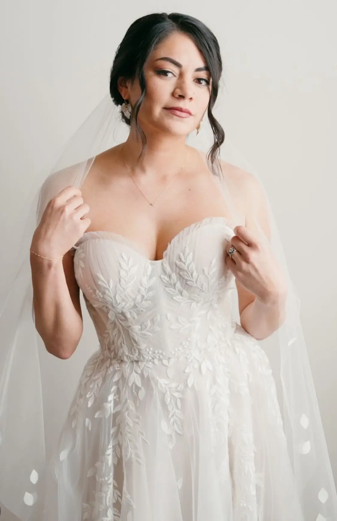 A woman wearing a strapless white wedding dress with leaf-patterned embroidery, holding her veil with both hands and looking at the camera. | Envy Studio, Lancaster, PA