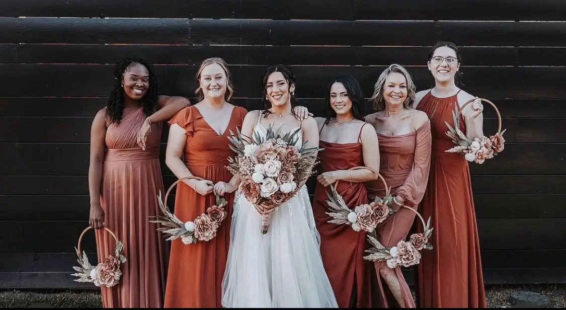 A bride in a white dress stands with five bridesmaids in rust-colored dresses, each holding a floral hoop, posed in front of a dark wooden wall. | Envy Studio, Lancaster, PA