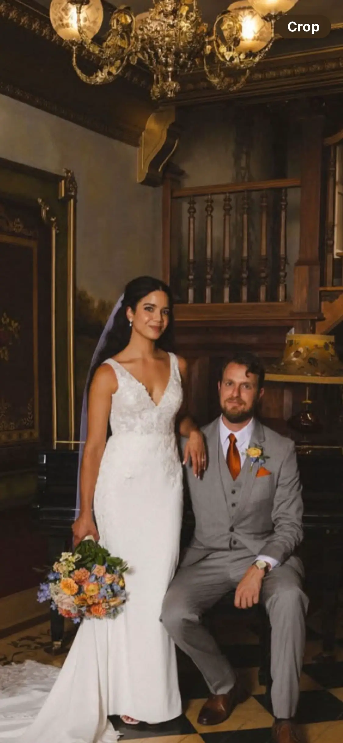 A bride in a white gown stands beside a seated groom in a gray suit with orange accents, posing in an ornate, vintage-style room. | Envy Studio, Lancaster, PA