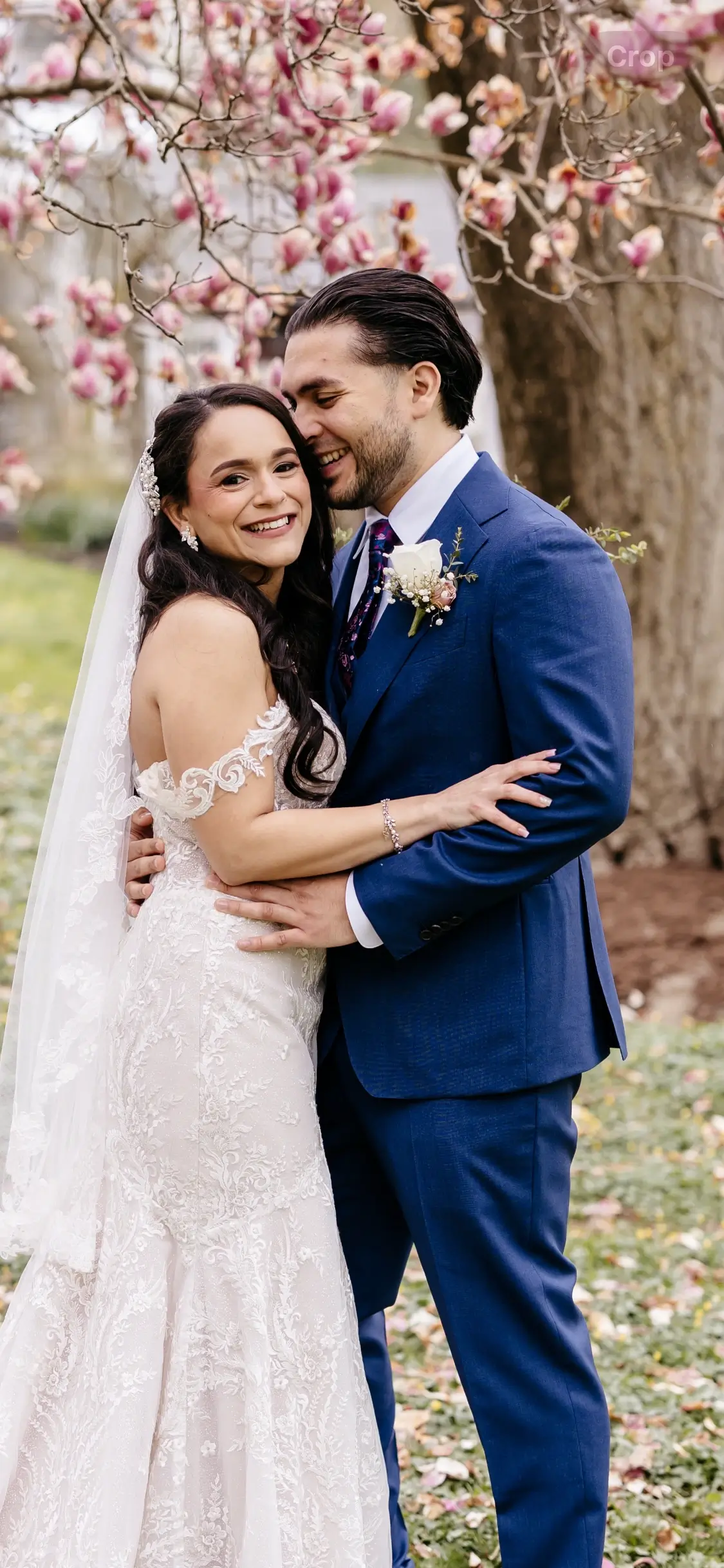 A bride and groom embrace outdoors under blooming trees, both smiling. The bride wears a white lace dress and veil; the groom is in a blue suit with a boutonniere. | Envy Studio, Lancaster, PA