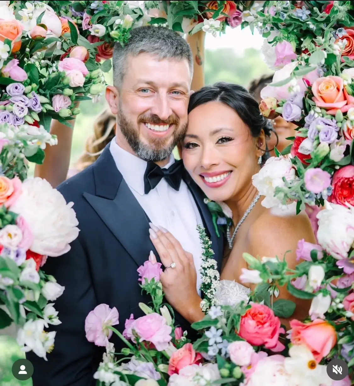 A smiling bride and groom pose closely together, framed by a vibrant floral arrangement featuring assorted colorful flowers. | Envy Studio, Lancaster, PA