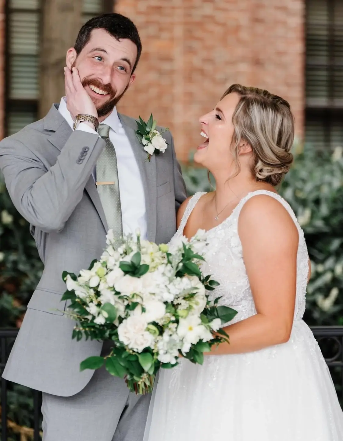 A bride in a white dress holding a bouquet smiles at a groom in a gray suit, who is touching his face and smiling. They are standing outdoors near greenery and a brick wall. | Envy Studio, Lancaster, PA
