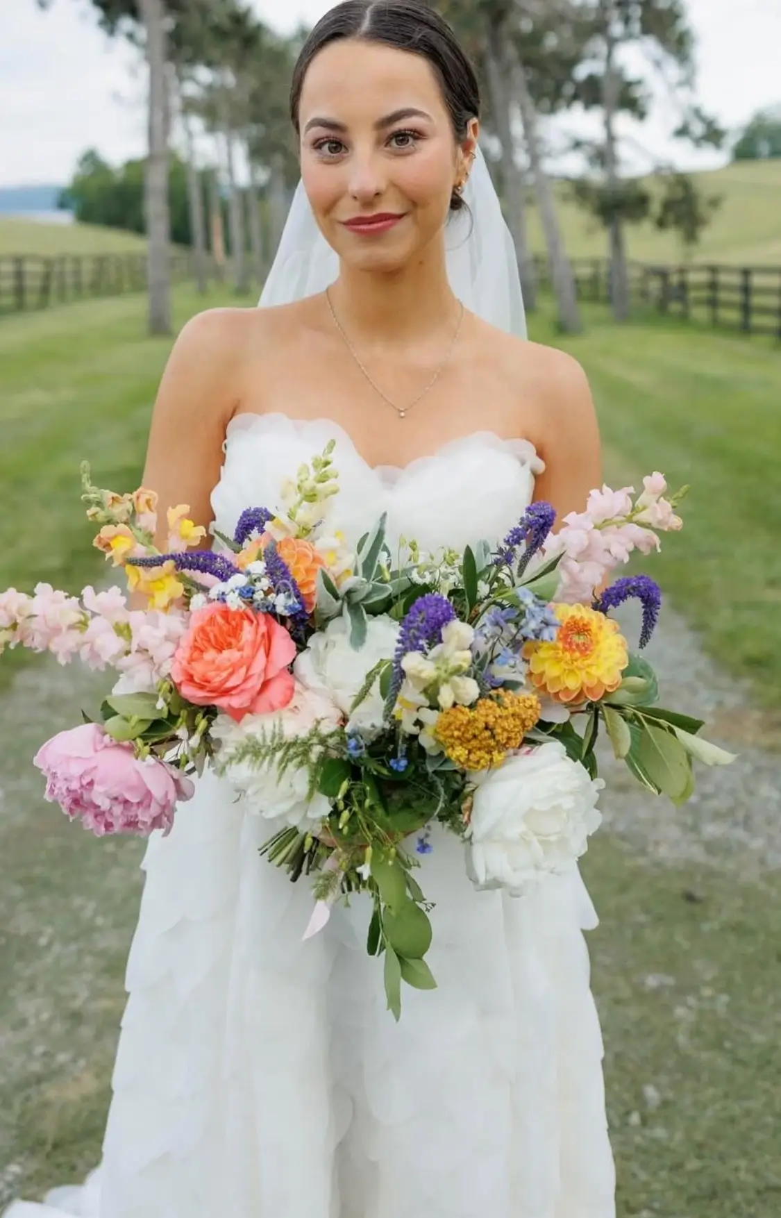 A bride in a strapless white wedding dress holds a large, colorful bouquet of flowers while standing outdoors on a grassy path. | Envy Studio, Lancaster, PA