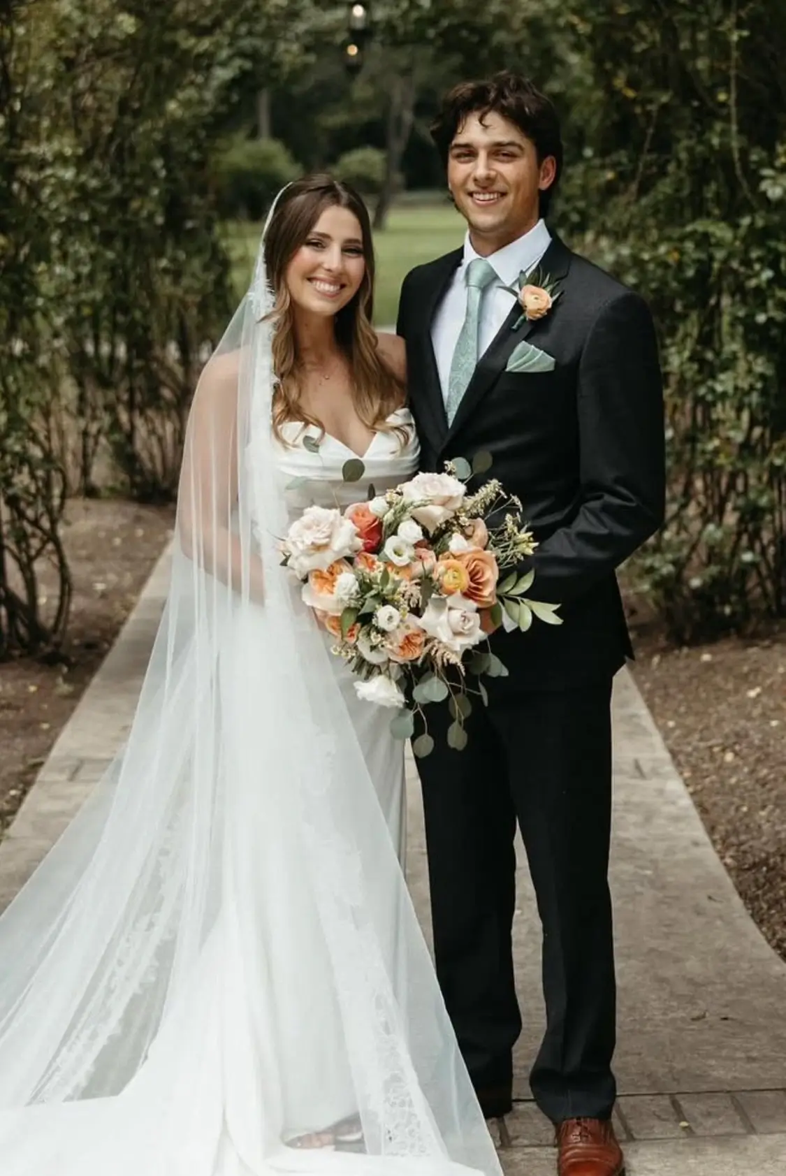 A bride in a white gown and veil stands next to a groom in a dark suit and green tie, holding a bouquet of flowers, posing outdoors on a garden path. | Envy Studio, Lancaster, PA