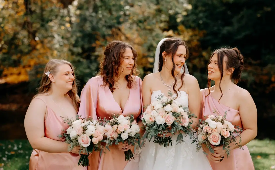 Four women, including a bride in white and three bridesmaids in pink dresses, stand outdoors holding bouquets of flowers and smiling at each other. | Envy Studio, Lancaster, PA