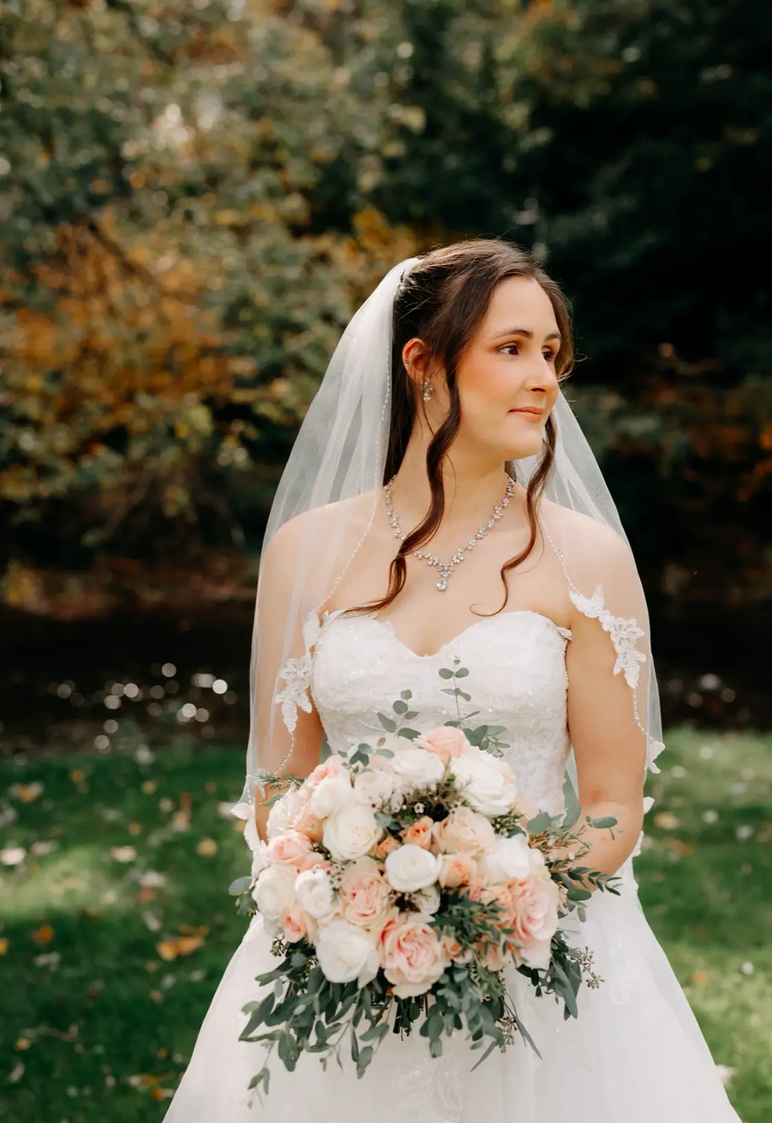 A bride in a white dress and veil holds a bouquet of flowers while standing outdoors in front of trees with autumn foliage. | Envy Studio, Lancaster, PA