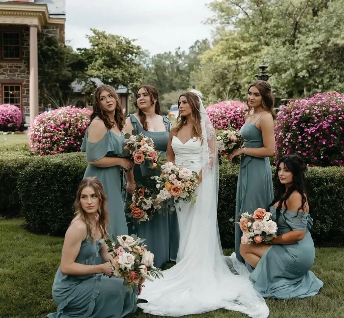 A bride in a white dress and veil stands outdoors with five bridesmaids in matching blue dresses, all holding bouquets, surrounded by greenery and blooming flowers. | Envy Studio, Lancaster, PA