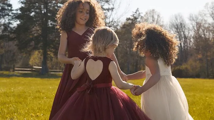 Three young girls in dresses hold hands and play in a grassy outdoor area with trees and sunlight in the background. | Envy Studio, Lancaster, PA