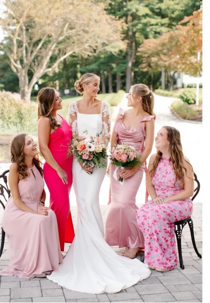 A bride in a white gown holds a bouquet and stands with four bridesmaids in pink dresses, posing outdoors on a paved area with trees in the background. | Envy Studio, Lancaster, PA