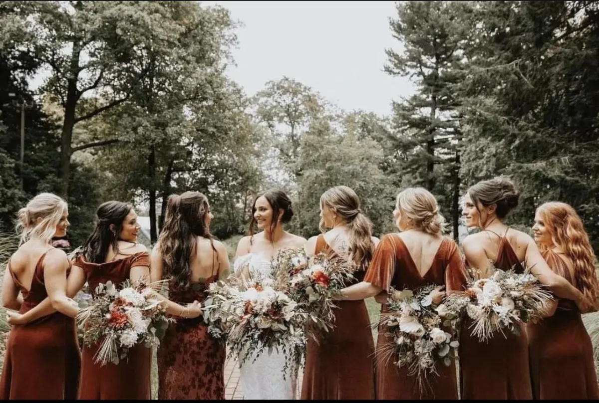 A bride in white stands outdoors with six bridesmaids in matching rust-colored dresses, all holding bouquets and facing away from the camera. Trees fill the background. | Envy Studio, Lancaster, PA