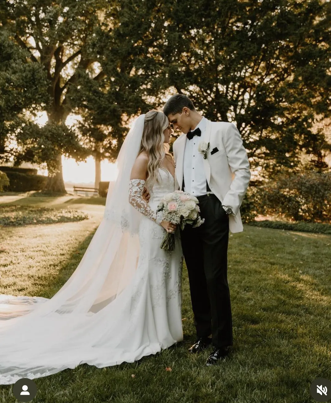 A bride and groom stand close together on grass, facing each other. The bride holds a bouquet and wears a long veil; the groom wears a white jacket and black pants. Trees are in the background. | Envy Studio, Lancaster, PA