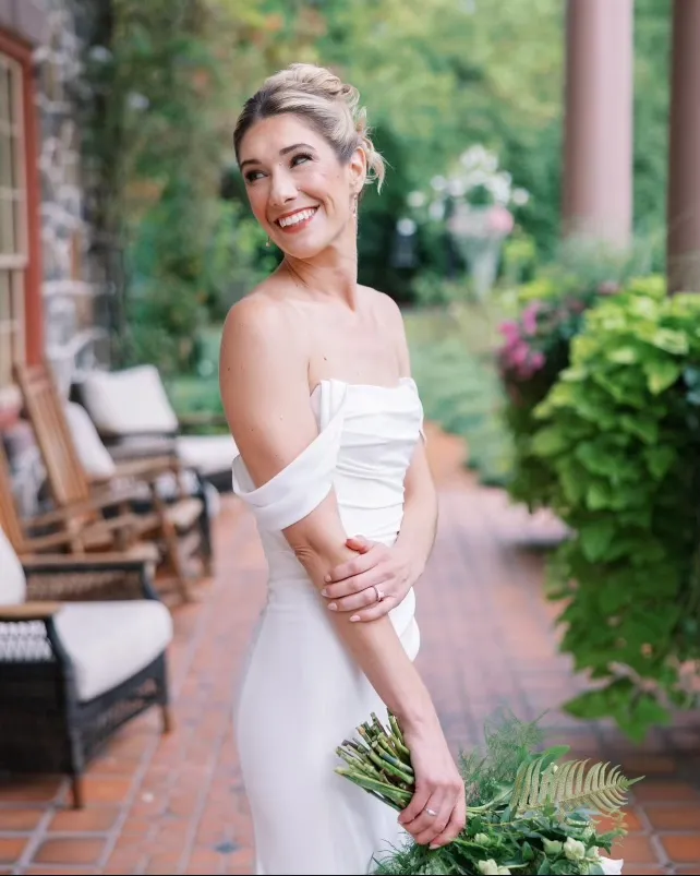 A woman in an off-the-shoulder white dress stands on a brick patio holding a bouquet, smiling with greenery and outdoor furniture in the background. | Envy Studio, Lancaster, PA