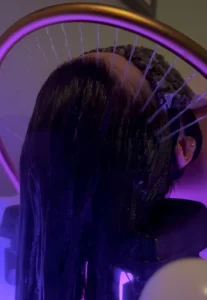 A person with dark hair is reclining at a salon sink, having their hair washed, with water spraying onto their scalp and purple lighting visible. | Envy Studio, Lancaster, PA