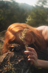 A person with long red hair leans on a fallen tree trunk, holding a twig with moss, with a forest background in soft focus. | Envy Studio, Lancaster, PA