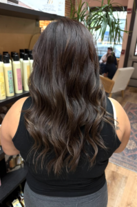 A person with long, wavy dark brown hair stands indoors near shelves of hair products at a Davines salon Lancaster PA, facing away from the camera. | Envy Studio, Lancaster, PA